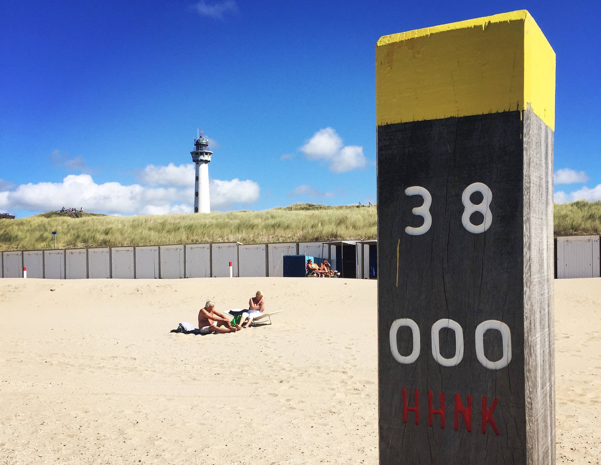 Strandpaal met gele bovenkant, op de achtergrond strandhuisjes en de vuurtoren Van Speijk